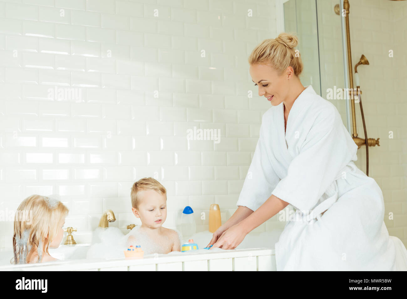 Glückliche junge Mutter in Bademantel spielen mit entzückenden kleinen Zicklein auf Badewanne sitzen Stockfoto
