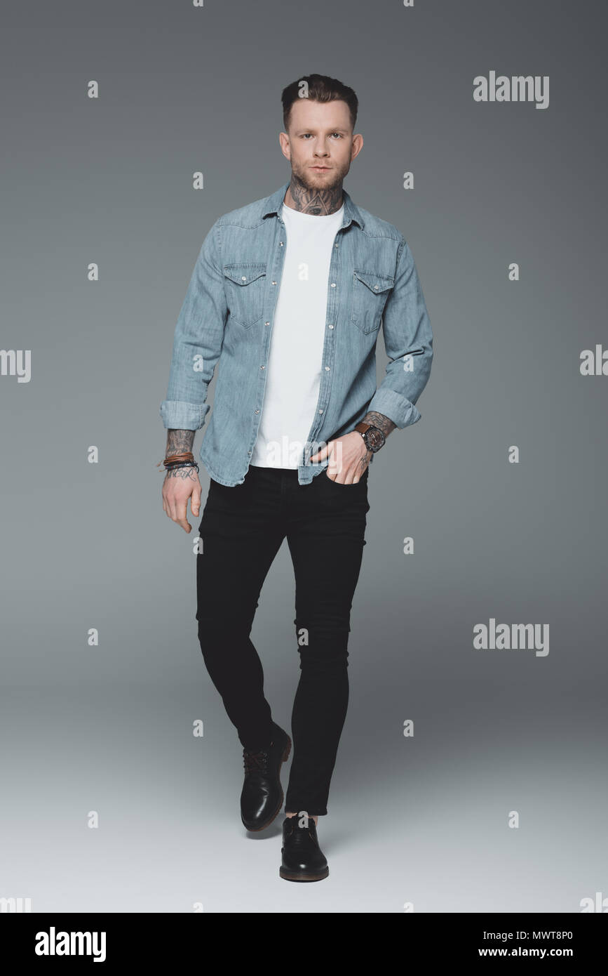 Volle Länge von stattlichen jungen tätowierten Mann mit der Hand in der Tasche und Kamera auf Grau Stockfoto