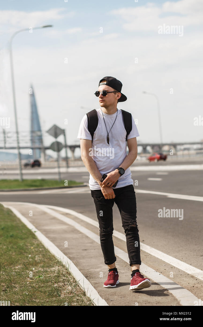 Stilvolle Mann per Anhalter auf der Straße oder Autobahn. Er trug, schwarze Jeans, Schwarzes T-Shirt, schwarze Kappe mit runden Sonnenbrille. Stockfoto