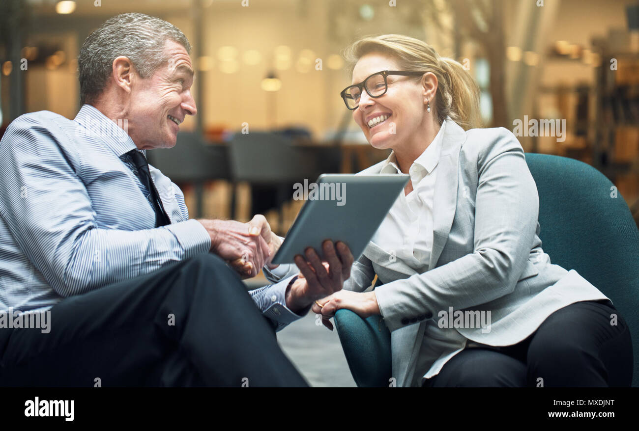 Lächelnd reife Geschäftsfrau Händeschütteln mit einem männlichen Kollegen halten einen digitalen Tablet beim Sitzen auf Stühlen in einem Büro Lobby Stockfoto