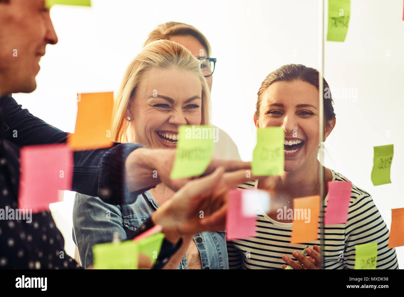 Lachend Gruppe von verschiedenen jungen Geschäftsleuten zusammen Brainstorming mit Haftnotizen auf eine Glaswand, während in einem modernen Büro stehend Stockfoto