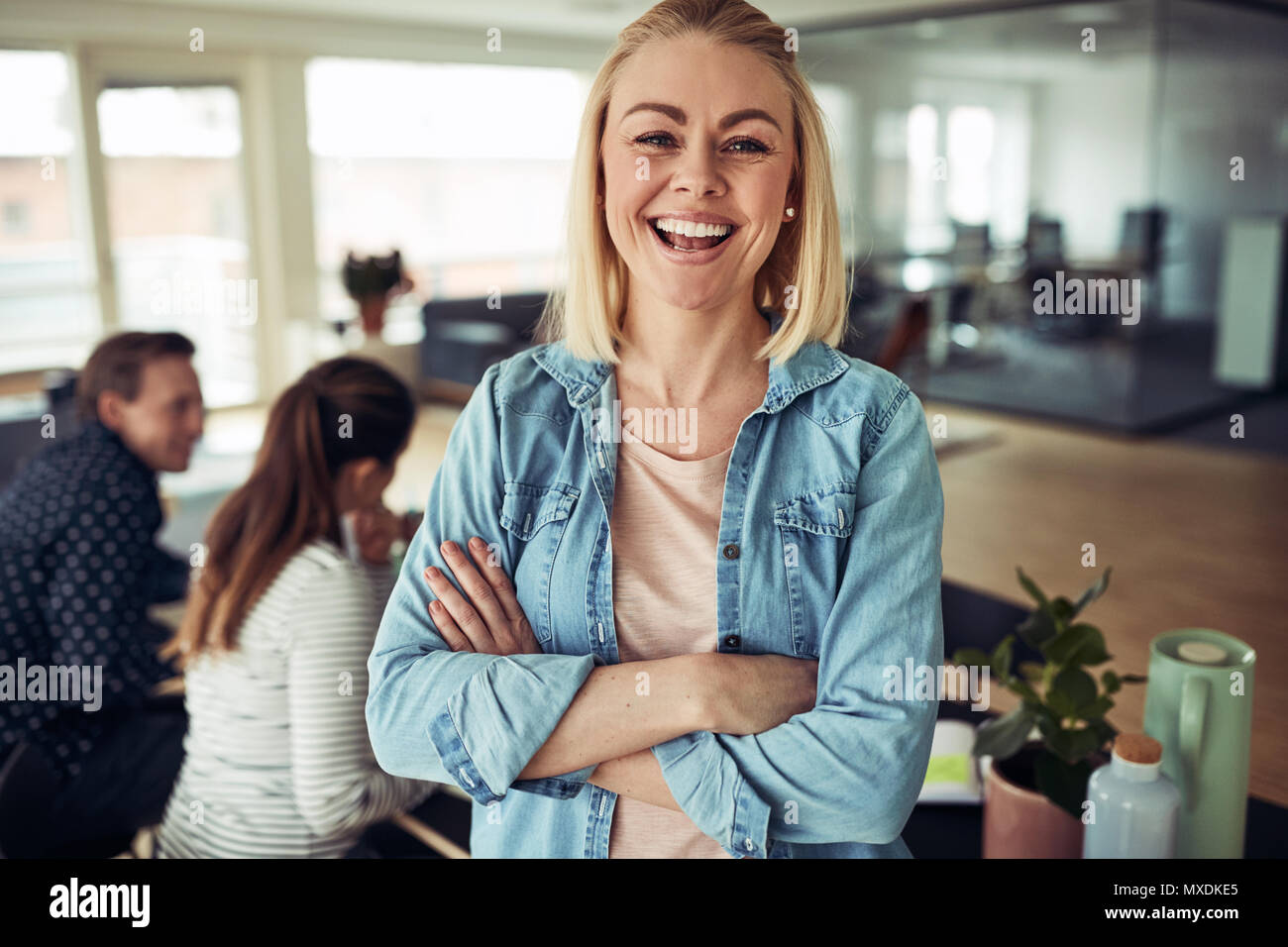 Lachende junge geschäftsfrau stehend, die Arme in einem Büro mit Kollegen an einem Tisch im Hintergrund arbeiten überschritten Stockfoto