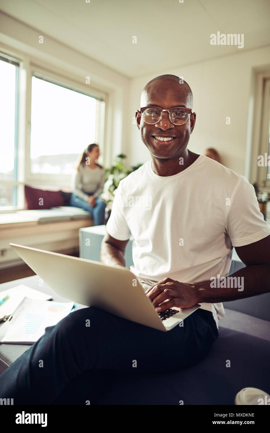 Lächelnd afrikanischen Geschäftsmann an seinem Schreibtisch in einem Büro an einem Notebook arbeitet mit Kollegen im Hintergrund sitzen Sitzen Stockfoto
