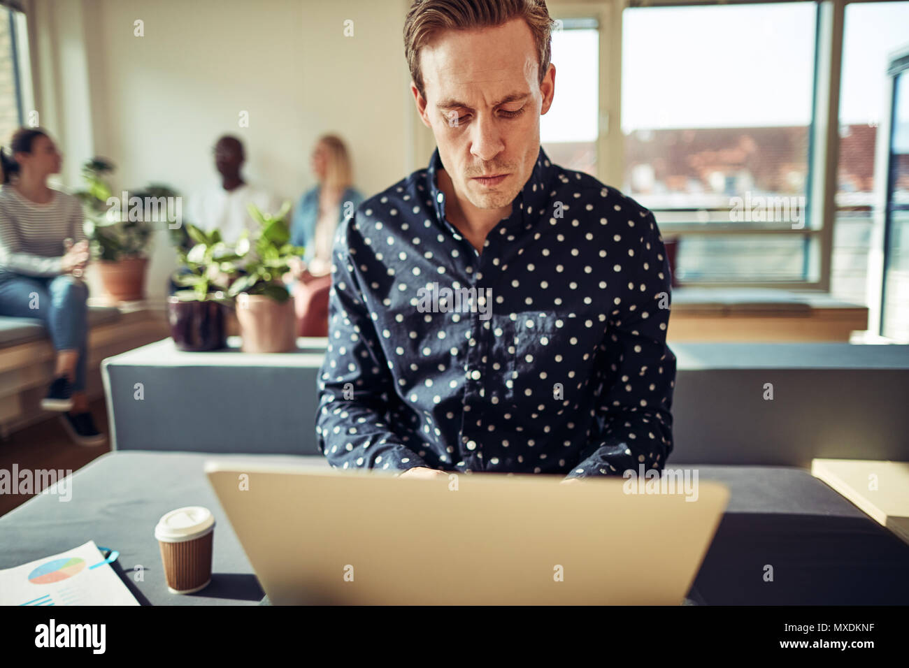 Konzentrierte sich junge Unternehmer online arbeiten mit einem Laptop sitzend auf seinem Schreibtisch in einem Büro mit Kollegen zusammen gesprochen im Hintergrund Stockfoto