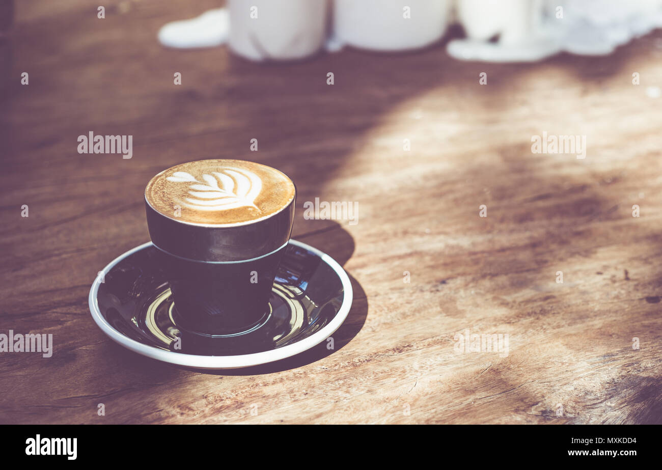 Close up Hot schwarz Kaffee Tasse mit Baum Herzform latte Art Milch Schaum auf Holz Tisch am Fenster mit harten Schatten Sonnenlicht im Cafe Restaurant. Stockfoto