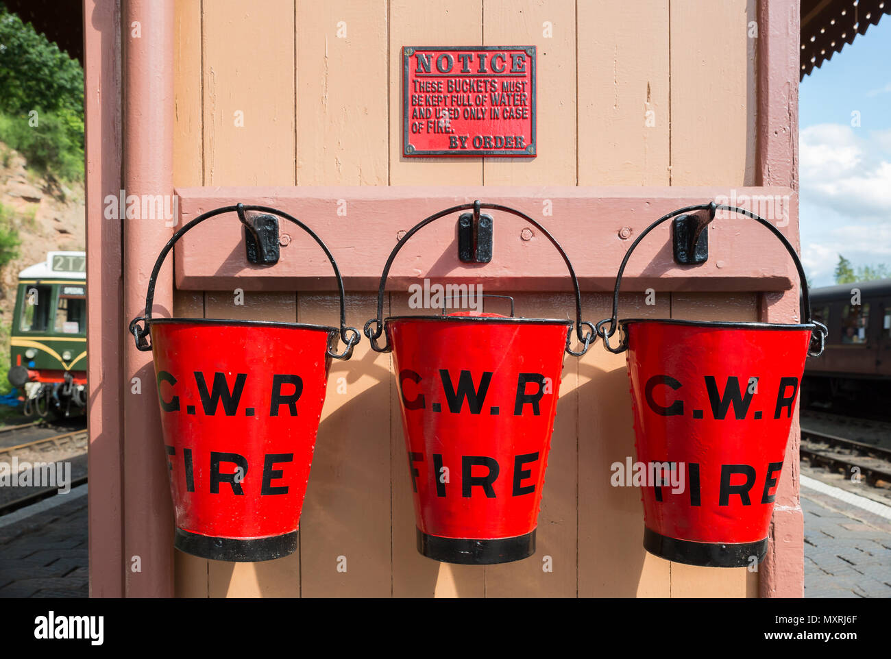 Nahaufnahme von drei roten GWR Notfeuerlöschpumpe Eimer auf Plattform im Vintage Steam Railway Station. Drei rote Eimer in einer Reihe. Trio der Eimer in der Sonne. Stockfoto