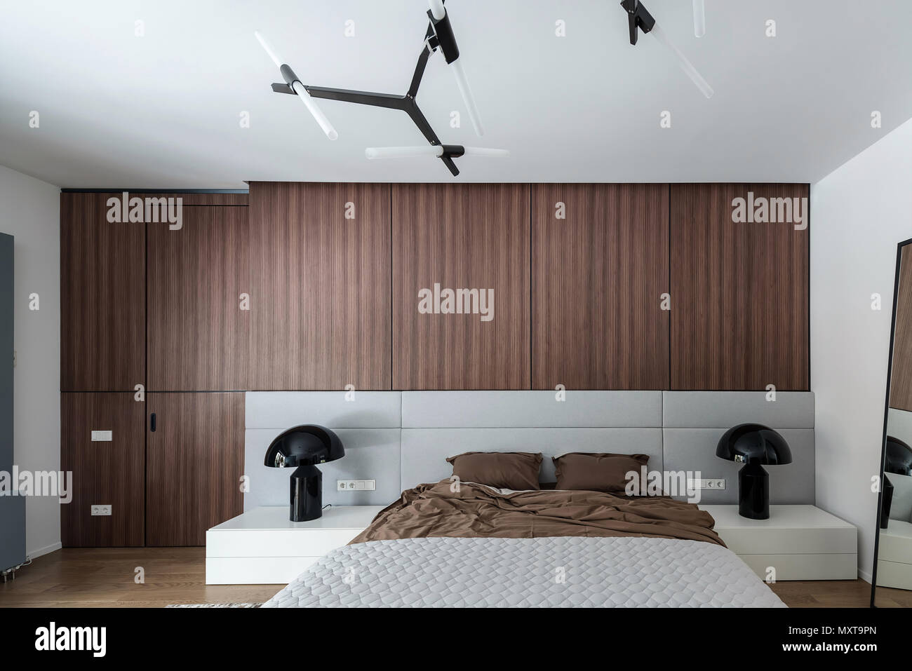 Modernes Schlafzimmer mit weißen Wänden und einem Parkett auf dem Fußboden. Es gibt ein Bett mit brauner Bettwäsche und einem Grau karierte, Spiegel, schwarzem Lampen auf steht, Stockfoto