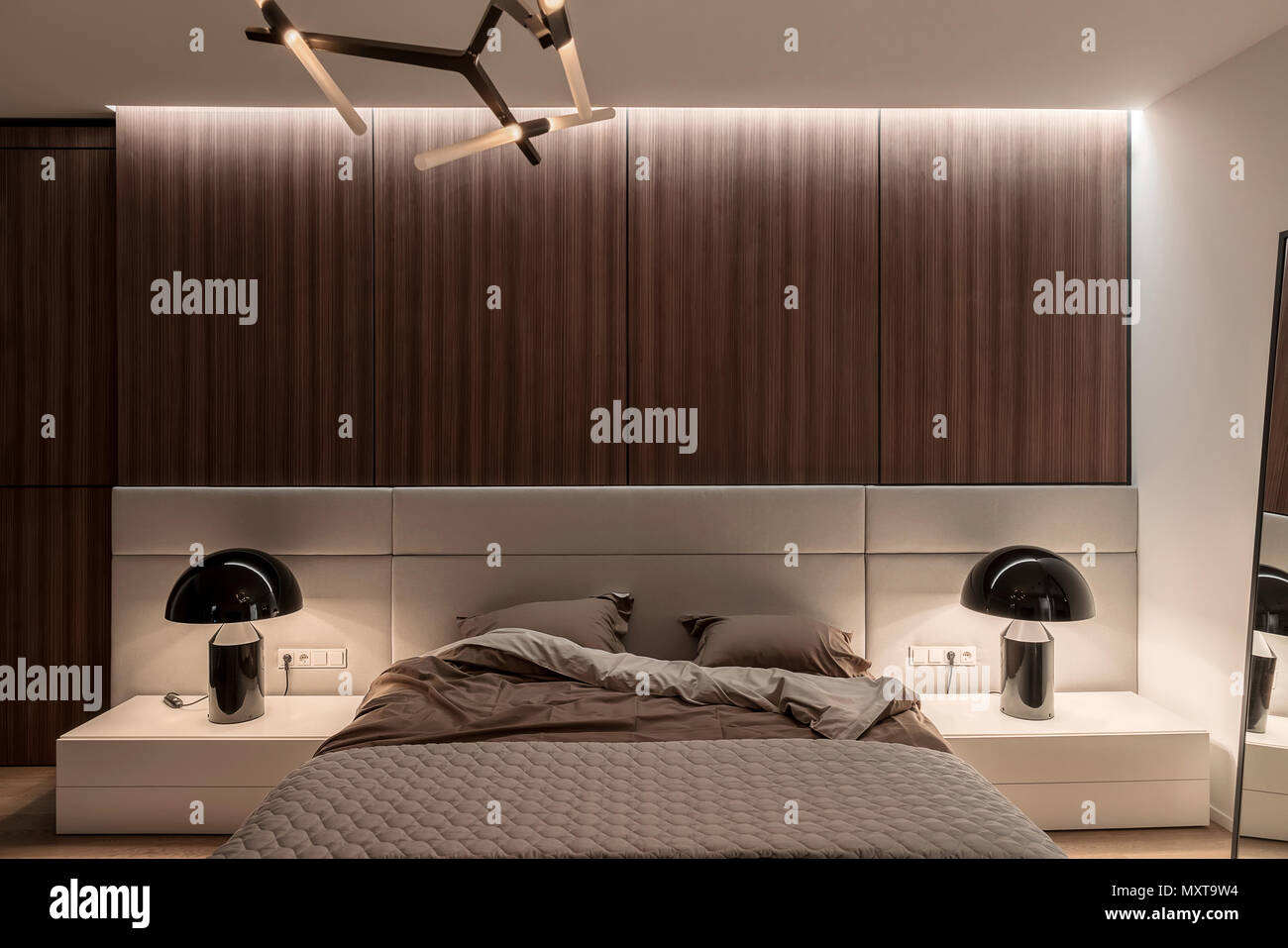 Schlafzimmer in einem modernen Stil mit weißen Wänden und Holzböden, Parkett auf dem Fußboden und helle fancy Lampen. Es gibt ein Bett mit brauner Bettwäsche und einem grauen plai Stockfoto