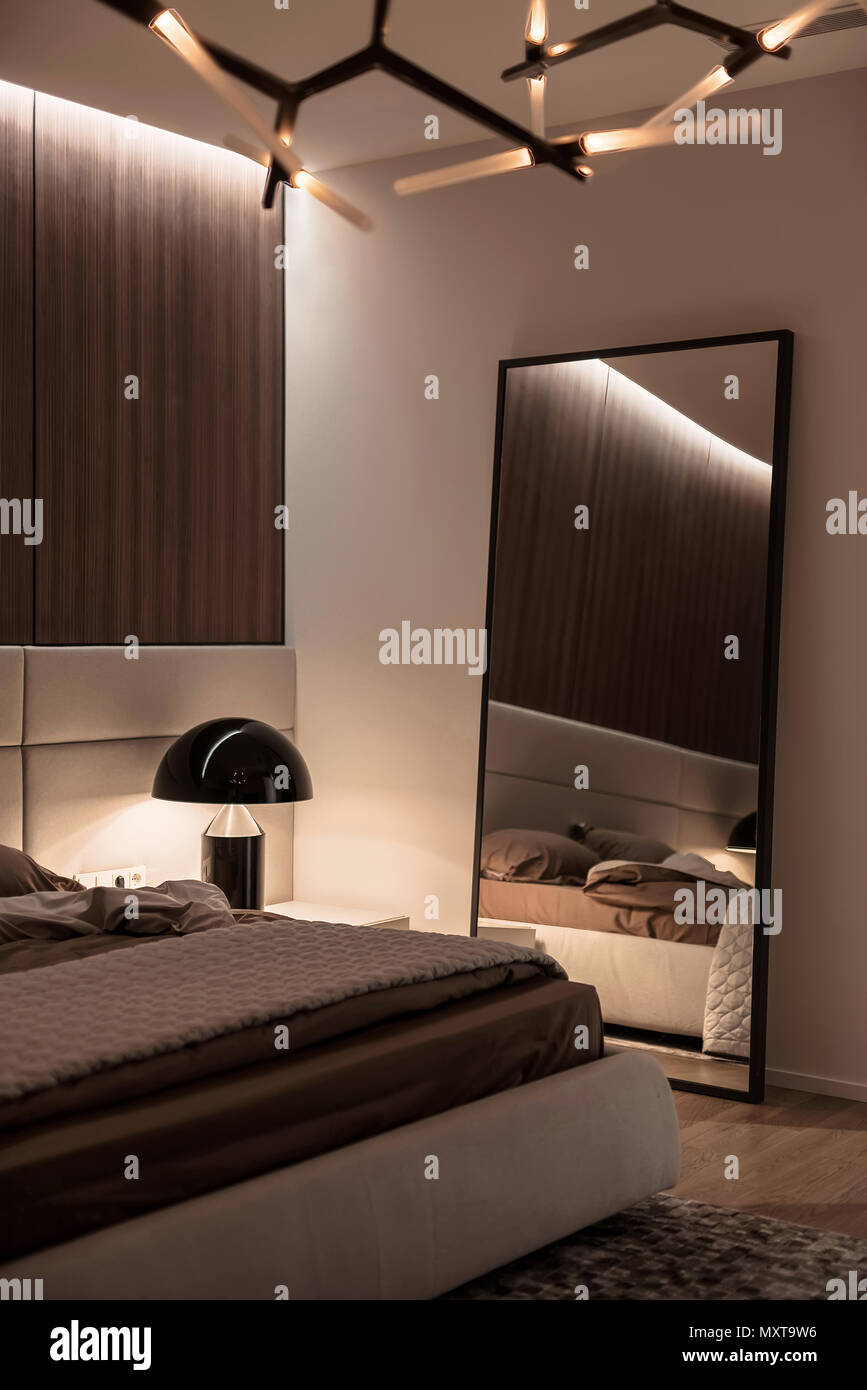 Modernes Schlafzimmer mit weißen Wänden und Holzböden, Parkett mit einem Teppich auf dem Boden und leuchtende Phantasie Lampen. Es gibt ein Bett mit brauner Bettwäsche und einem Grau p Stockfoto