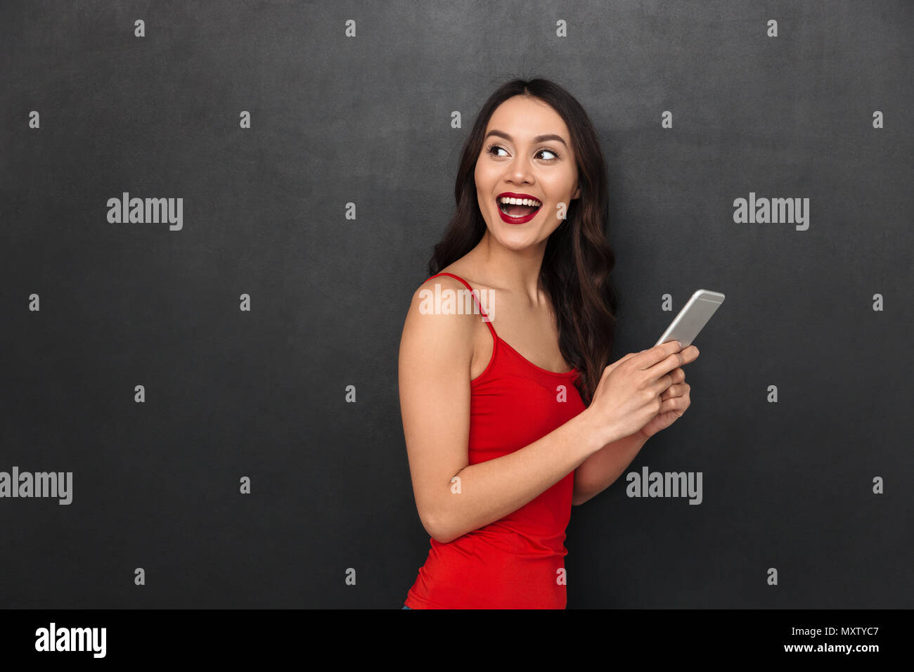 Bild der nette junge asiatische Frau stehend über dunkle graue Wand Hintergrund zur Seite schauen, Chatten per Smartphone. Stockfoto