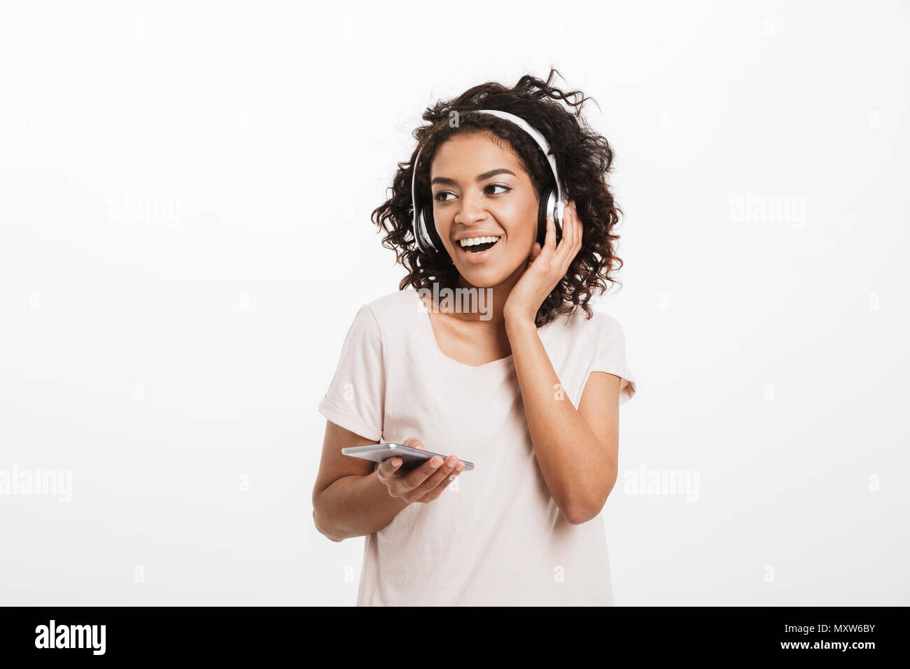 Moderne amerikanische Frau mit Afro Frisur und Lächeln Hören von Musik über drahtlose Kopfhörer und Holding Handy in der Hand über Weiß isoliert Stockfoto