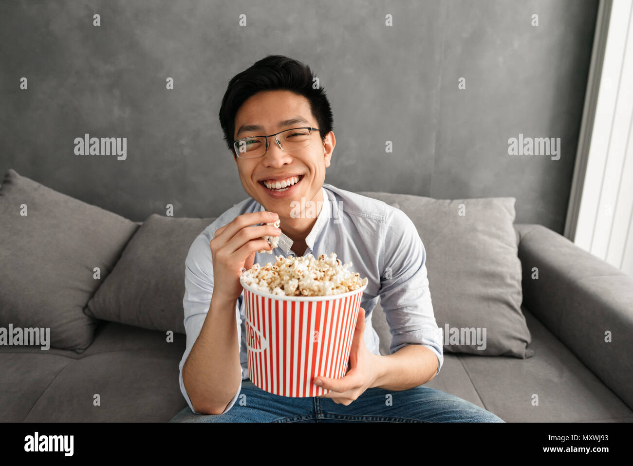 Porträt einer fröhlicher junger asiatischer Mann essen Popcorn während auf der Couch zu Hause sitzen und Fernsehen Stockfoto