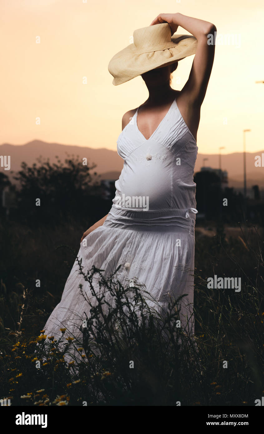 Schwangere Frau tragen weiße Kleid und Hut posiert im Freien Stockfoto