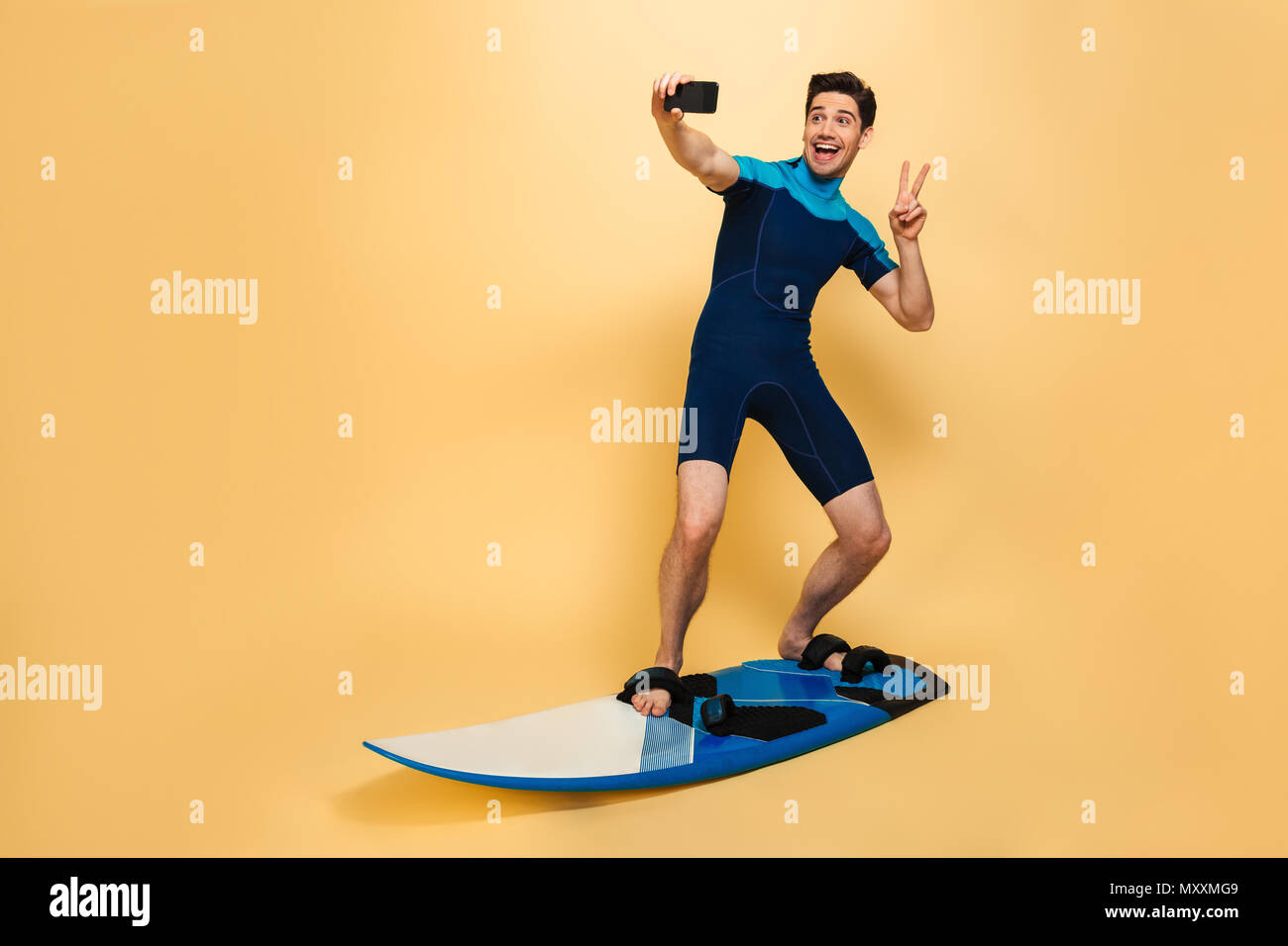 In voller Länge Porträt der glückliche junge Mann in Badeanzug ein selfie gekleidet beim Surfen auf einem Brett über Gelb Hintergrund isoliert Stockfoto