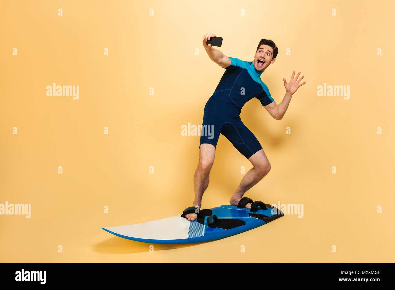In voller Länge Porträt von einem Aufgeregten jungen Mann im Badeanzug ein selfie gekleidet beim Surfen auf einem Brett über Gelb Hintergrund isoliert Stockfoto