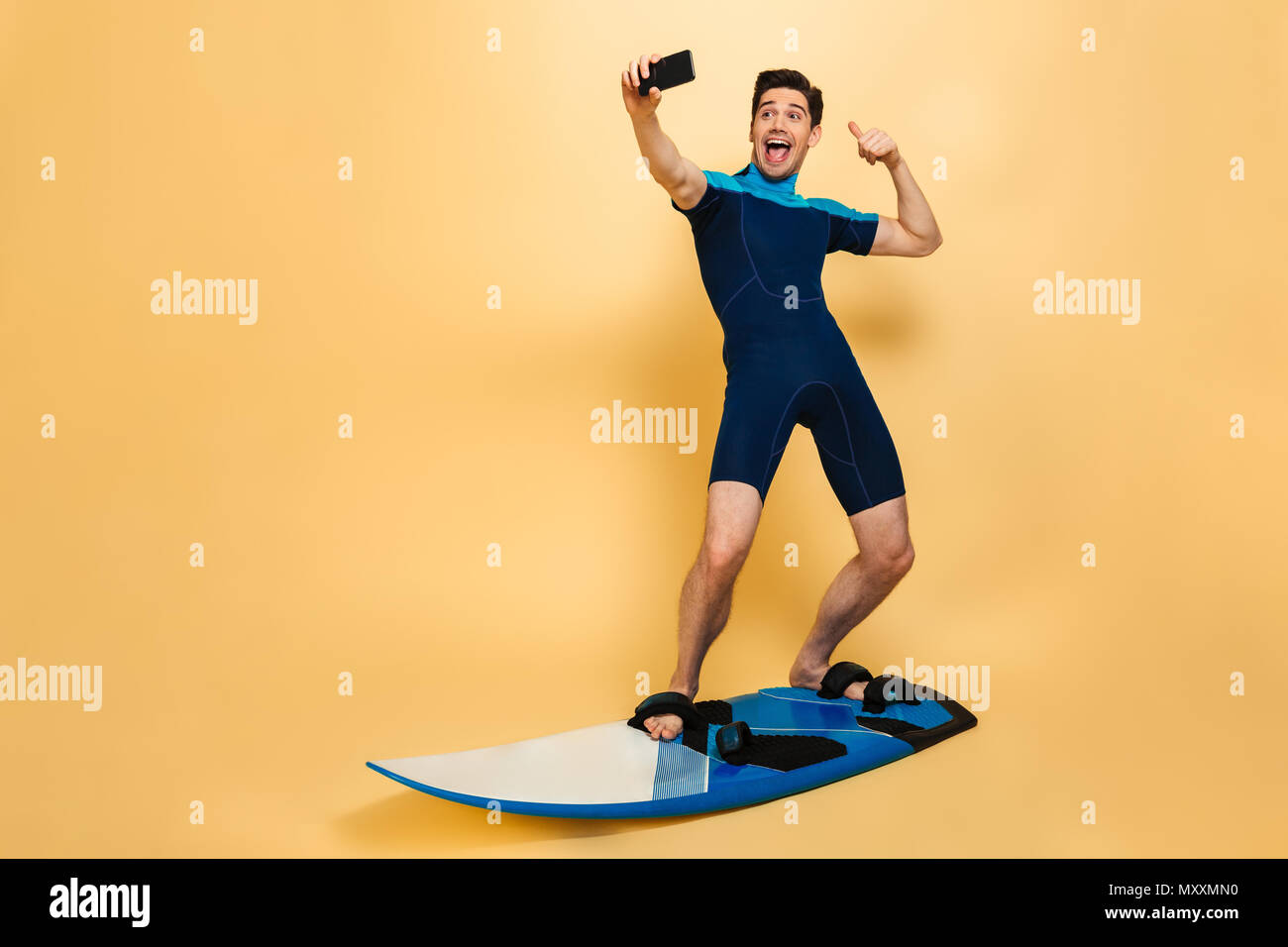 In voller Länge Porträt einer fröhlichen jungen Mann im Badeanzug ein selfie gekleidet beim Surfen auf einem Brett über Gelb Hintergrund isoliert Stockfoto