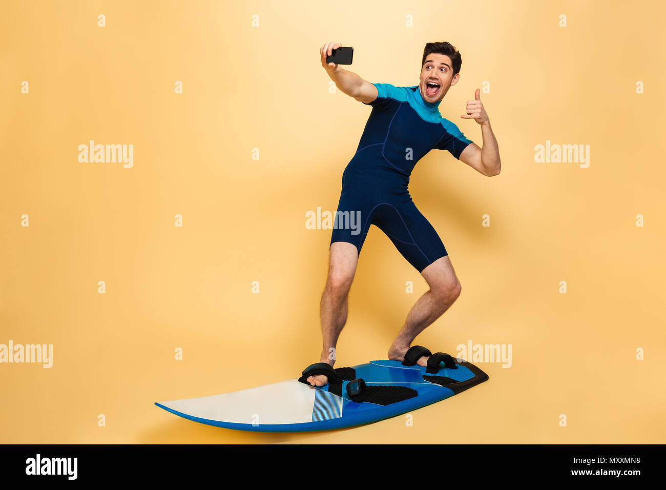 In voller Länge Porträt einer selbstbewussten jungen Mann im Badeanzug ein selfie gekleidet beim Surfen auf einem Brett über Gelb Hintergrund isoliert Stockfoto