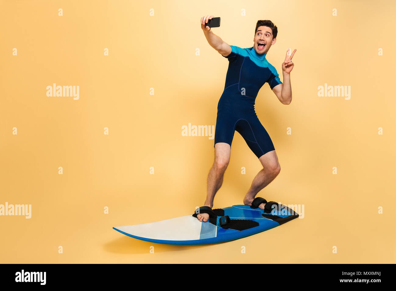 In voller Länge Porträt von einem freundlichen jungen Mann in Badeanzug ein selfie gekleidet beim Surfen auf einem Brett über Gelb Hintergrund isoliert Stockfoto