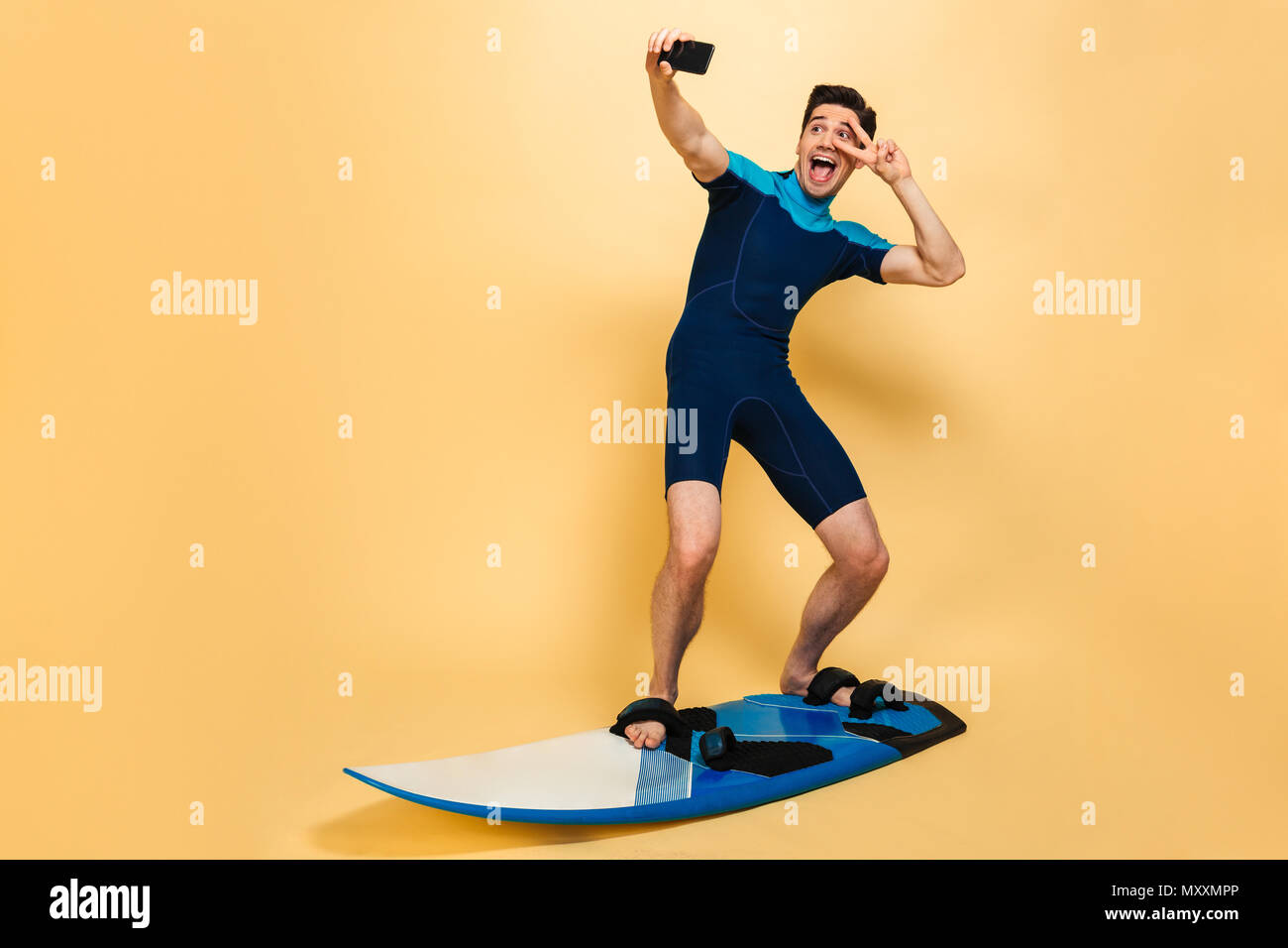 In voller Länge Porträt eines lächelnden jungen Mann im Badeanzug ein selfie gekleidet beim Surfen auf einem Brett über Gelb Hintergrund isoliert Stockfoto