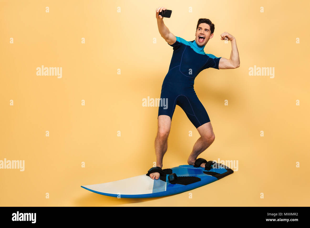 In voller Länge Porträt einer starken jungen Mann im Badeanzug ein selfie gekleidet beim Surfen auf einem Brett über Gelb Hintergrund isoliert Stockfoto