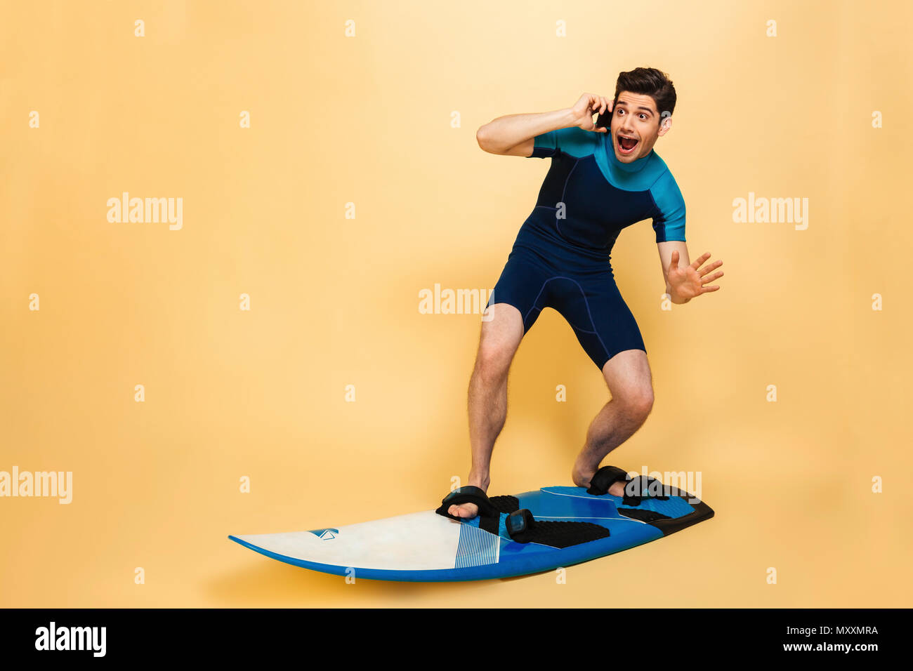 Foto von einem schreienden Angst junger Mann im Badeanzug auf gelbem Hintergrund auf Surfen board Gespräch per Telefon isoliert gekleidet. Stockfoto