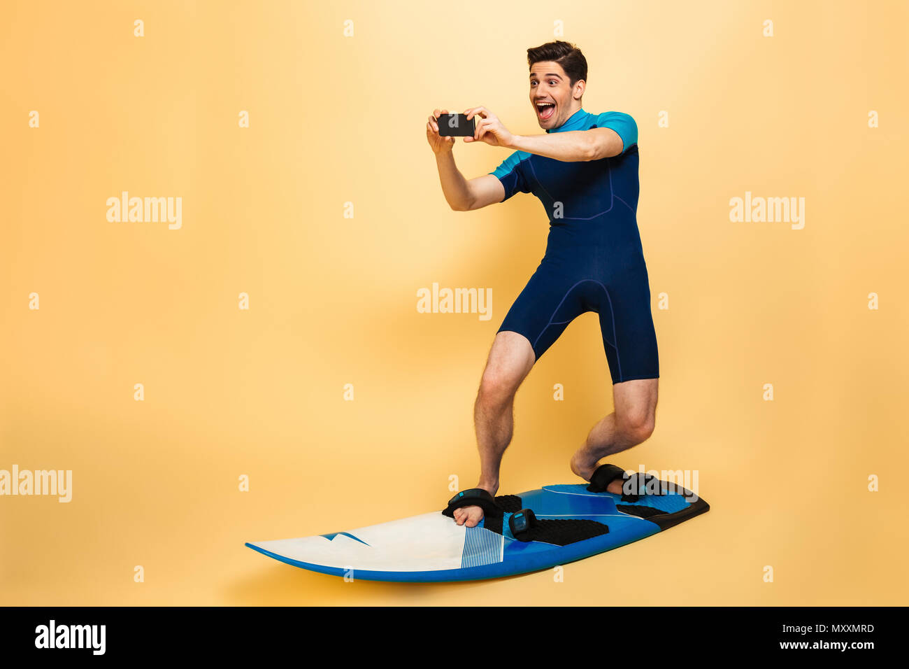 In voller Länge Porträt einer selbstbewussten jungen Mann in Badehose gekleidet sprechen ein Bild mit Handy beim Surfen auf einem Brett über Gelb ba isoliert Stockfoto