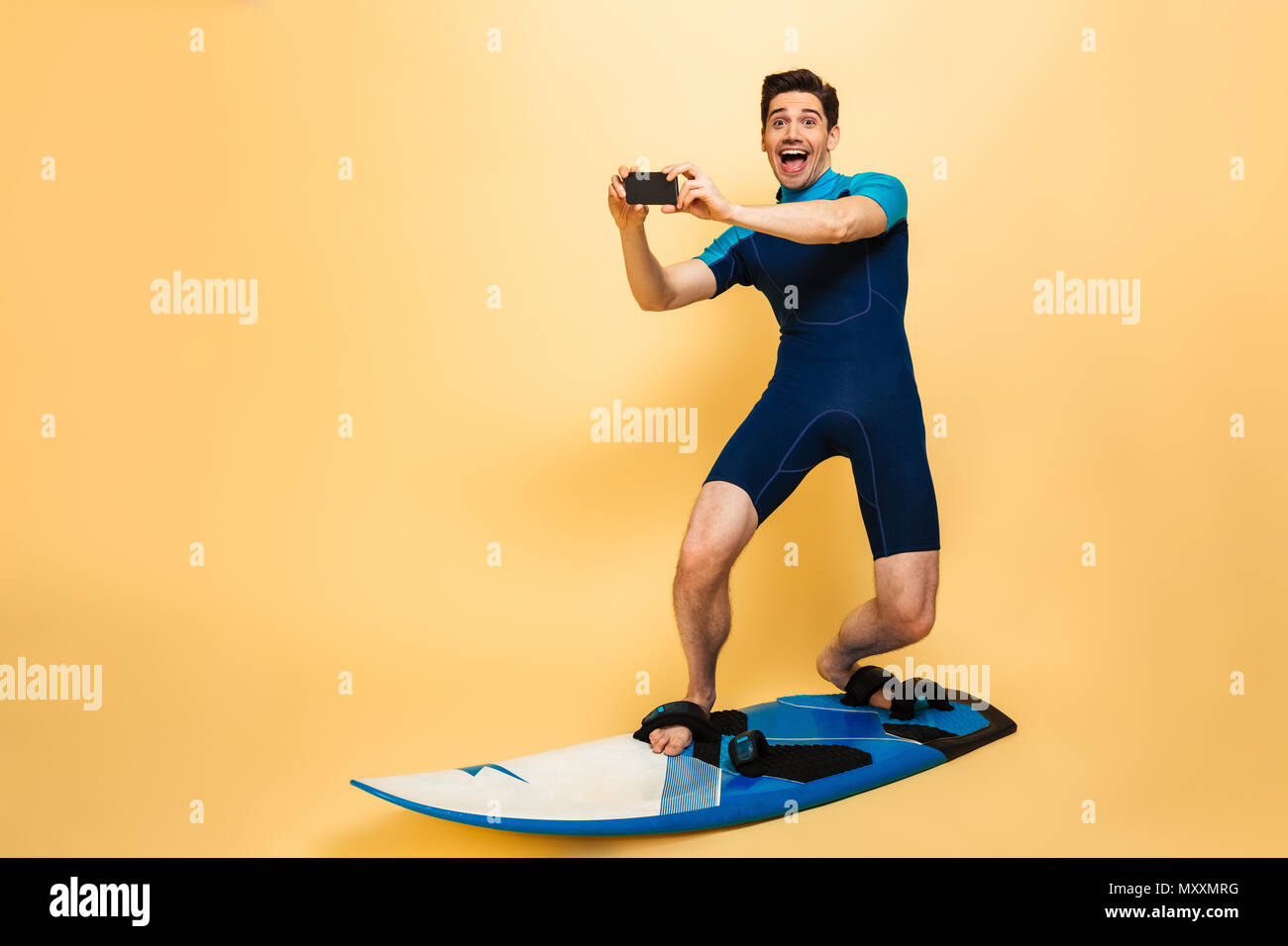 In voller Länge Porträt von einem Aufgeregten jungen Mann in Badeanzug bekleidet sprechen ein Bild mit Handy beim Surfen auf einem Brett über Gelb bac isoliert Stockfoto