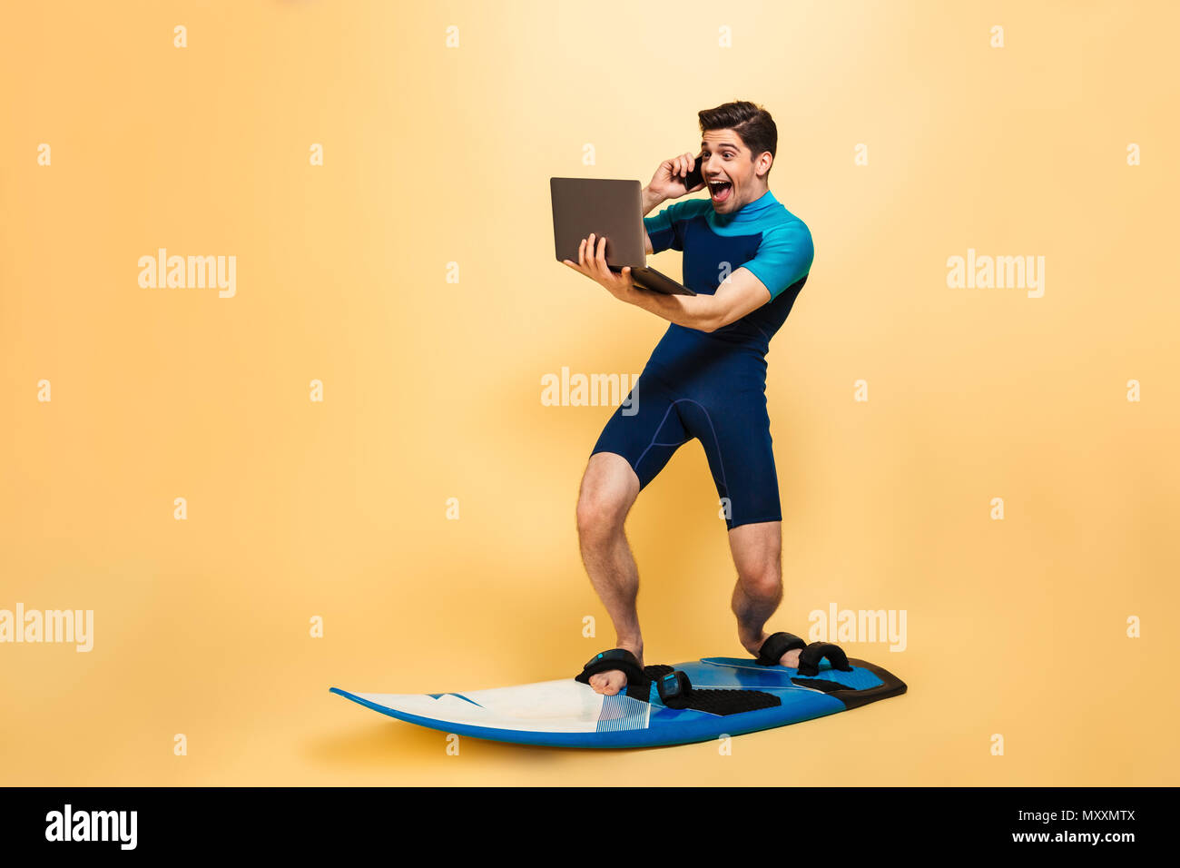 Foto einer aufgeregt, junger Mann im Badeanzug auf gelbem Hintergrund auf Surfen board Reden über Laptop Telefon isoliert gekleidet. Stockfoto