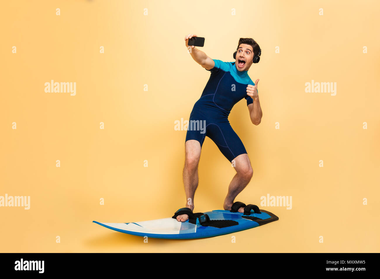 Foto von einem emotionalen junger Mann im Badeanzug auf gelben Hintergrund isoliert auf Surfen board selfie per Telefon hören Musik mit headpho gekleidet Stockfoto