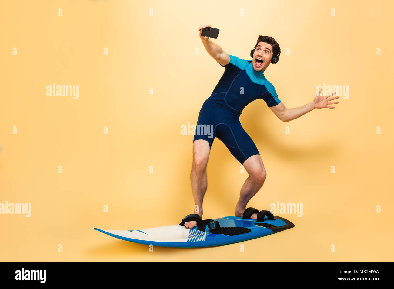 Foto des jungen Mannes im Badeanzug auf gelbem Hintergrund auf Surfen board selfie per Telefon Musik hören mit Kopfhörern isoliert gekleidet. Stockfoto