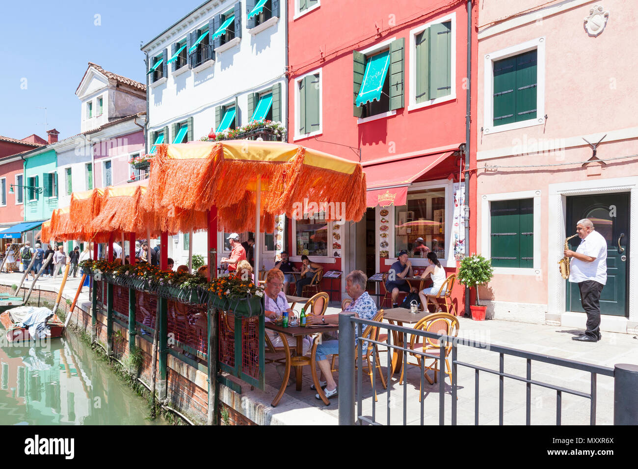 Menschen speisen zu den klassischen live Musik mit einem Saxophon Spieler in Insel Burano, Venedig, Venetien, Italien neben dem Kanal unter bunten orange Umbria Stockfoto
