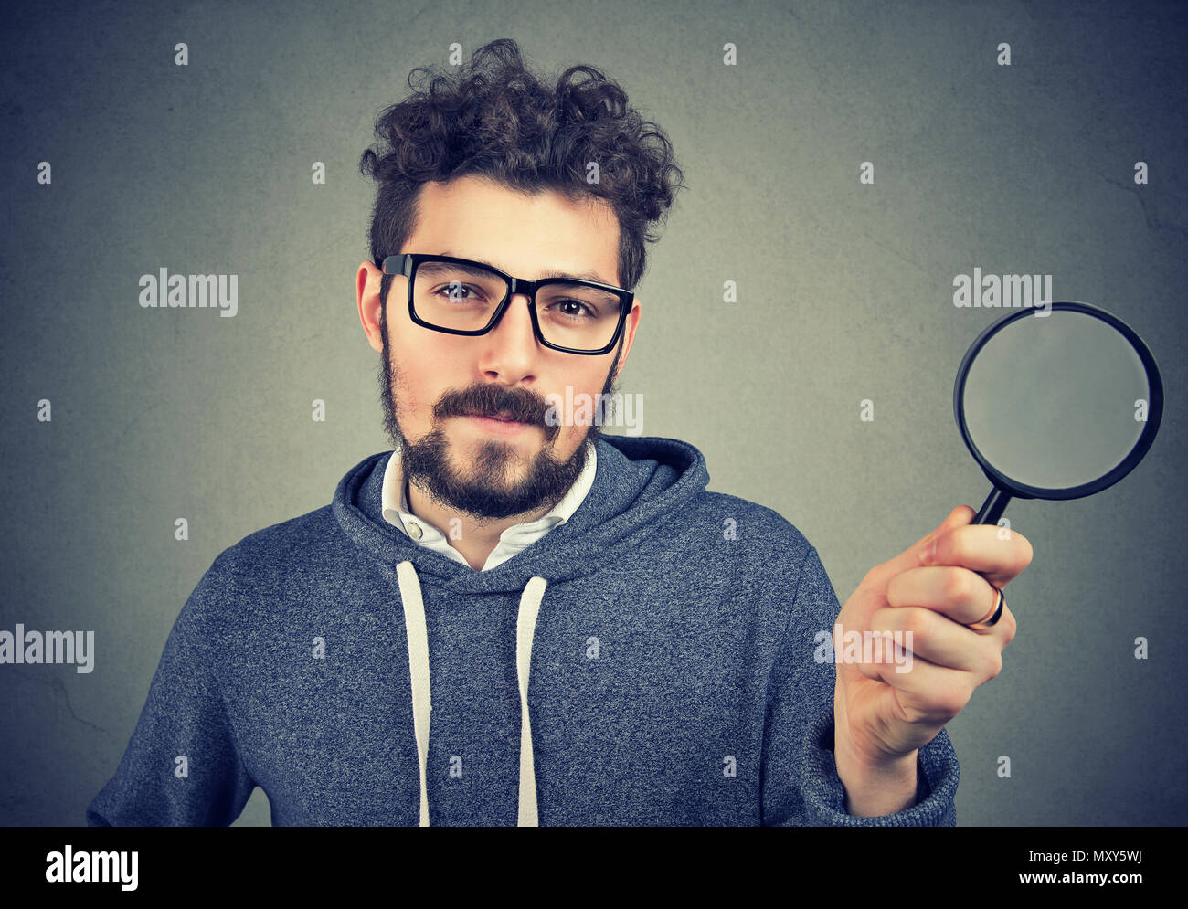 Bärtige gewissenhafter Mann in Brille mit Lupe Kamera schaut auf grauem Hintergrund. Stockfoto