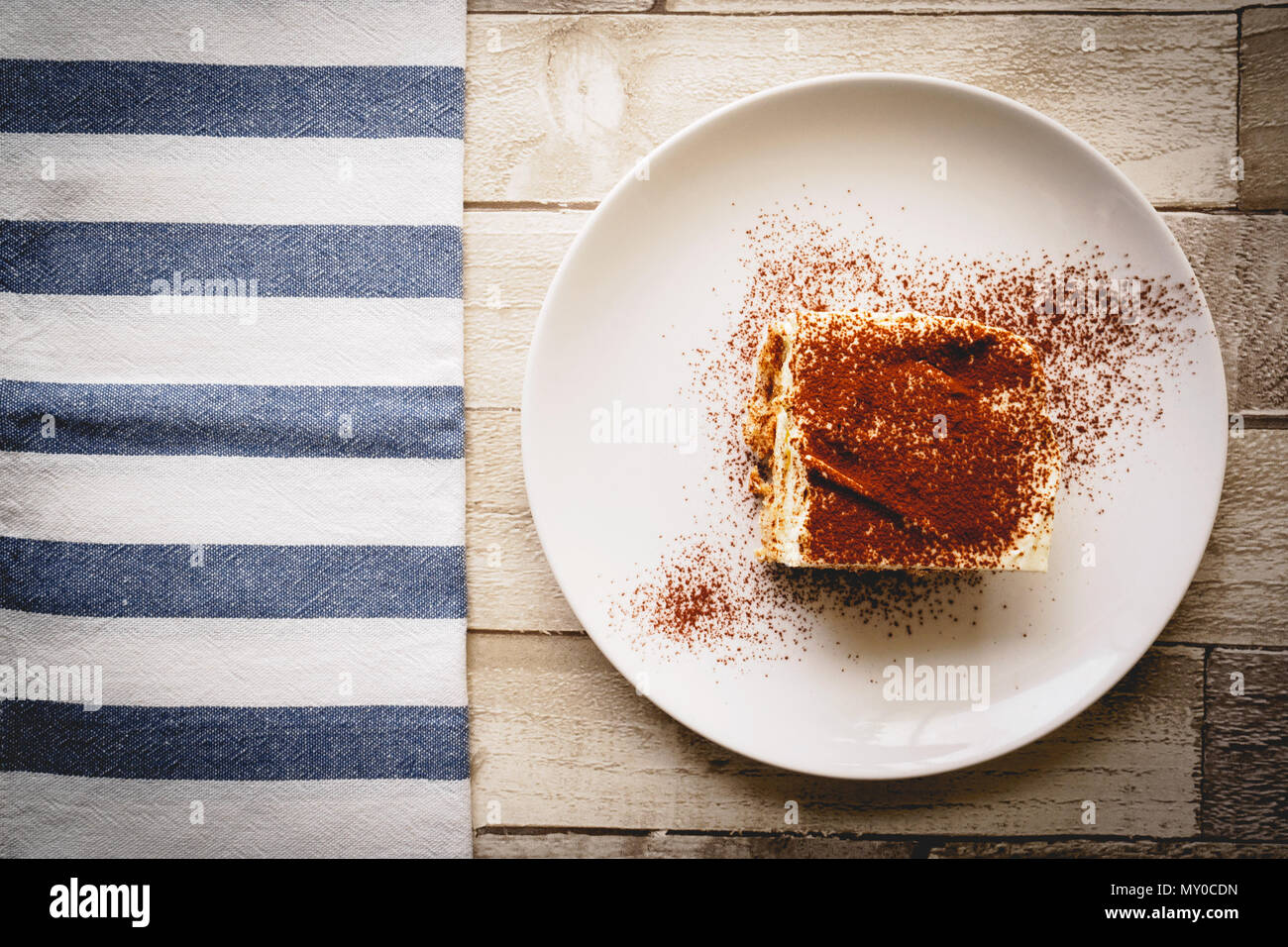 Ansicht von oben ein Stück hausgemachte Tiramisu (traditionelle italienische Dessert) auf einem weißen Teller auf einem Holzbrett mit einem weißen und blauen Küche Tuch. Stockfoto