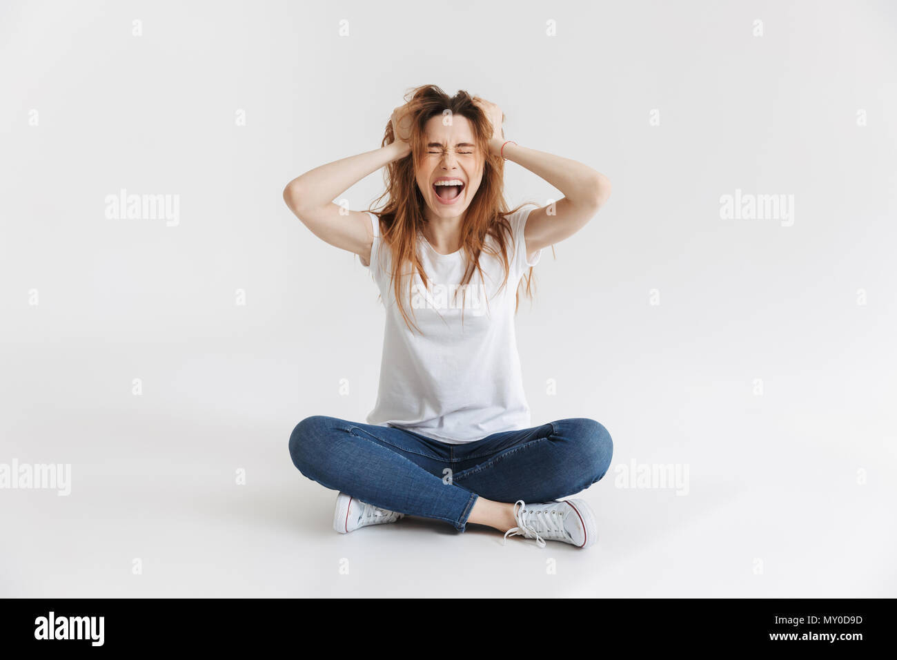 Wütende Frau im T-Shirt sitzen auf dem Boden und schrie mit geschlossenen Augen, während ihr Kopf über grauer Hintergrund Stockfoto