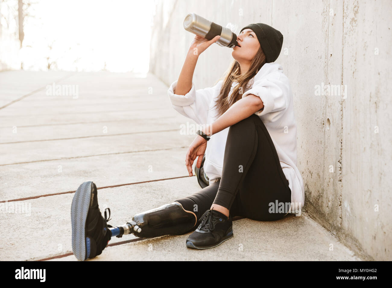 Foto von attraktiven jungen behinderten Frau, die bionischen Bein in Streetwear, sitzen auf den Betonboden outdoor und Trinkwasser aus Metall Schale Stockfoto