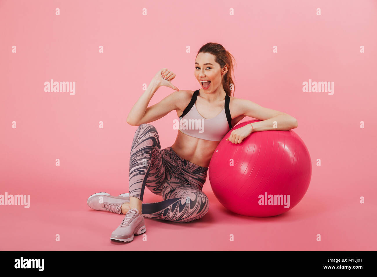 Gerne Sportlerin sitzen auf dem flloor mit Fitness Ball während Zeigt an sich, und wenn man die Kamera über rosa Hintergrund Stockfoto