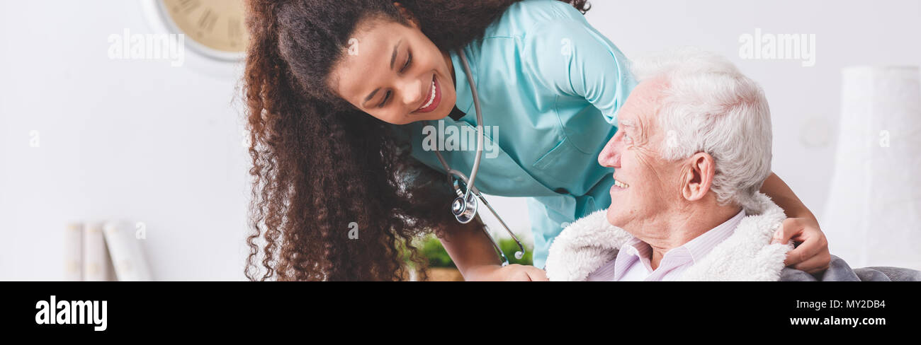 Panorama von einer glücklichen Krankenschwester mit einem Stethoskop für einen älteren Mann mit einer Decke in einem Pflegeheim Stockfoto