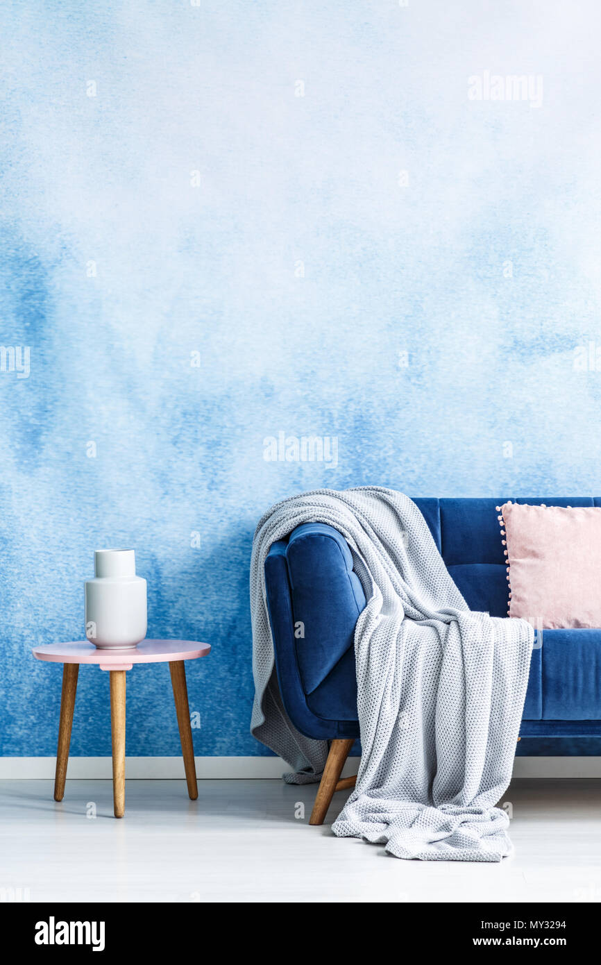 7/8-Foto von einer Couch mit einem grauen Decke neben einem Hocker mit einer weißen Vase in einem Wohnzimmer Interieur mit einer leeren Wand Stockfoto