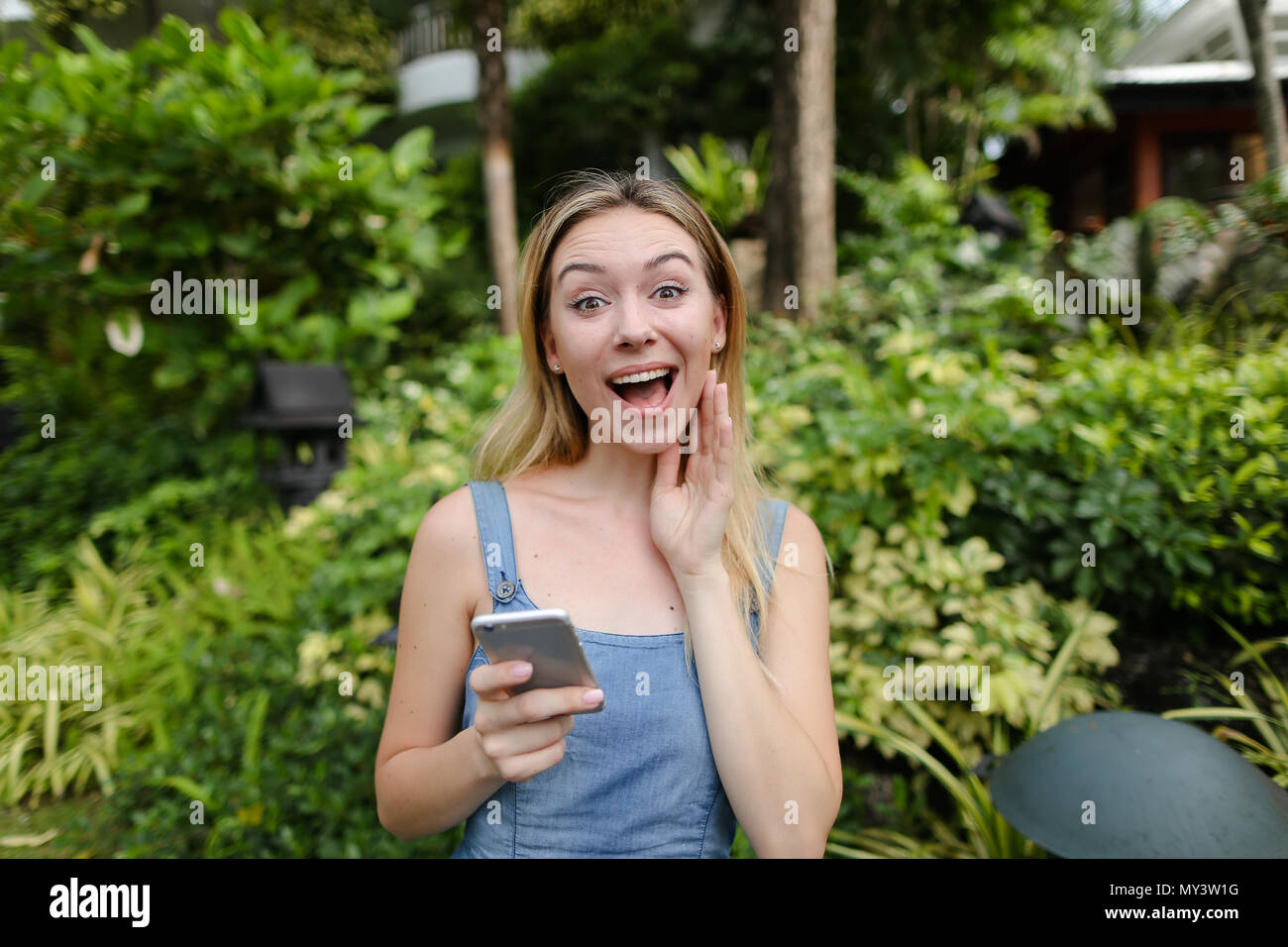 Junge jocund Frau sprechen, die von Smartphones im Garten in der Nähe von Bäumen, in Jeans sundress. Stockfoto