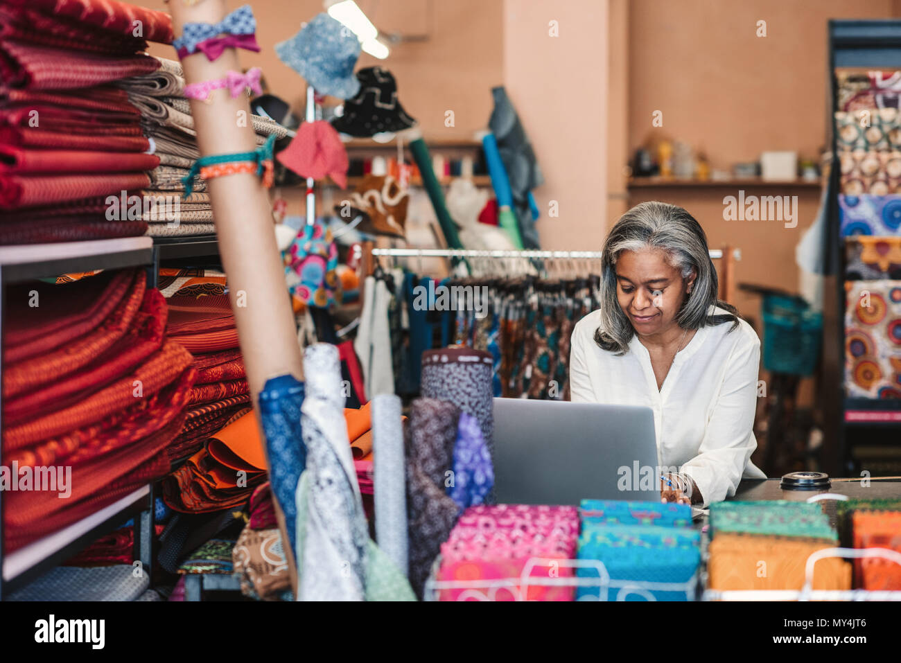Lächelnd reife Stoff shop besitzer, der an einem Notebook arbeitet, während bei einem Zähler von bunten Tüchern und Textilien umgeben stehend Stockfoto