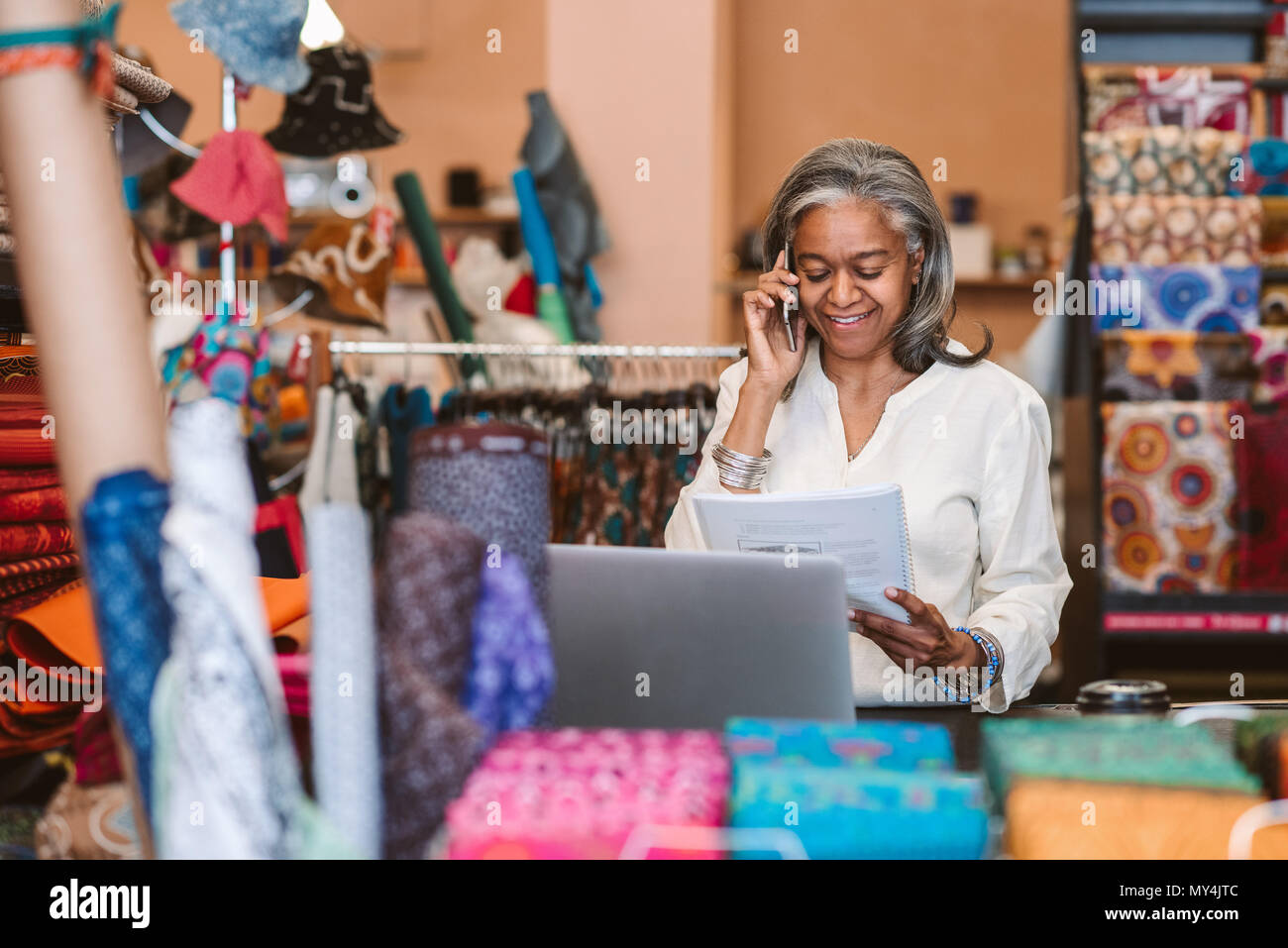 Lächelnd reife Stoff shop besitzer hinter einen Zähler von bunten Textilien Schreibarbeit Lesung und Gespräch am Mobiltelefon umgeben stehend Stockfoto
