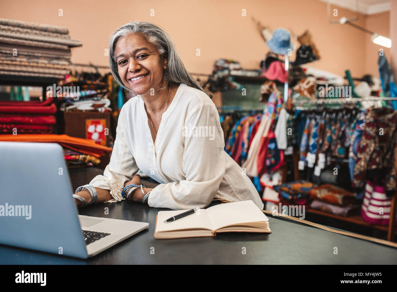Porträt eines lächelnden reife Stoff shop besitzer, der an einem Notebook arbeitet und schreiben Sie sich Notizen zu Ihren store Counter von bunten Textilien umgeben Stockfoto