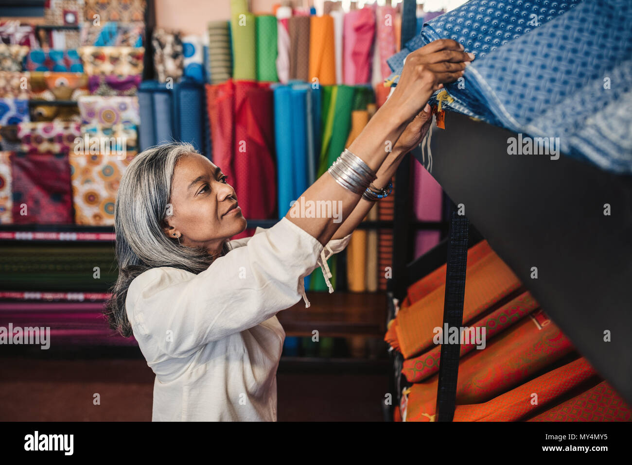 Lächelnd, reife Frau sucht durch die Rollen von bunten Textilien auf einem Regal beim Einkaufen in einem Stoff shop Stockfoto