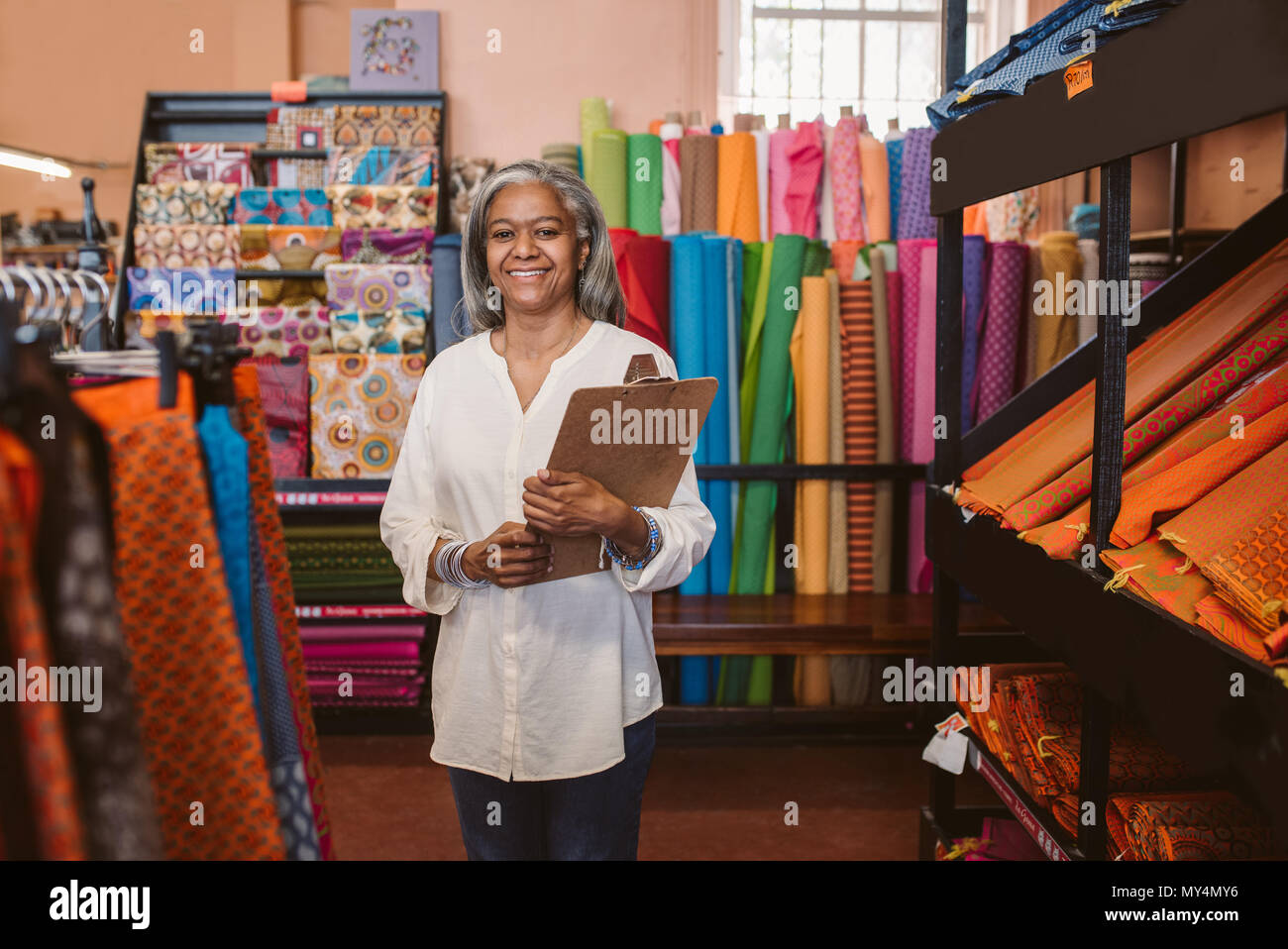 Porträt eines lächelnden reife Stoff shop besitzer eine Zwischenablage beim Stehen neben den Regalen mit bunten Tüchern und Textilien Stockfoto