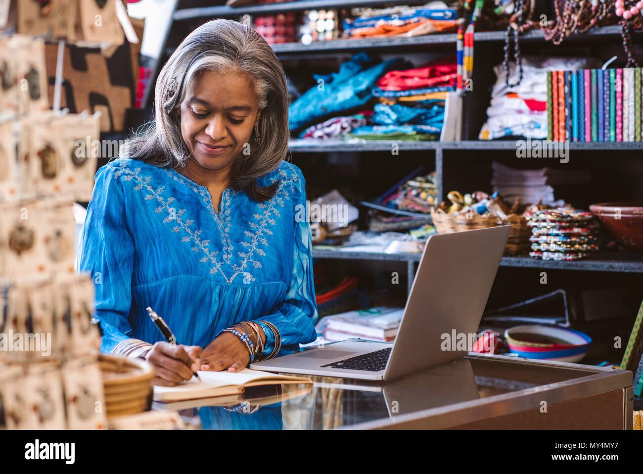 Lächelnd reife Stoff shop besitzer Schreiben in ein Notebook und arbeitet an einem Laptop auf einen Zähler von bunten Tüchern und Textilien umgeben Stockfoto