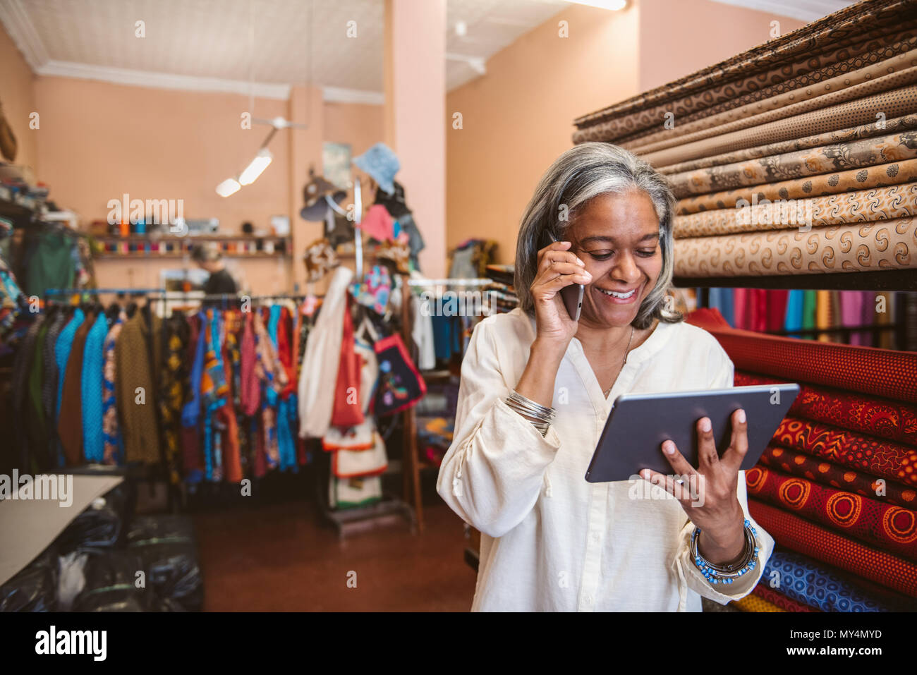 Lächelnd reife Stoff shop besitzer mit einem digitalen Tablet und Gespräche am Mobiltelefon im Stehen von bunten Tüchern und Textilien Stockfoto