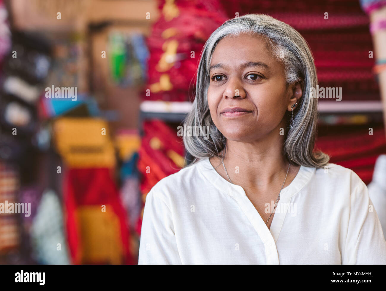 Reife weibliche Fabric store Inhaber stehen in Ihrem Shop mit Regalen der farbenfrohe Stoffe und Tücher im Hintergrund Stockfoto
