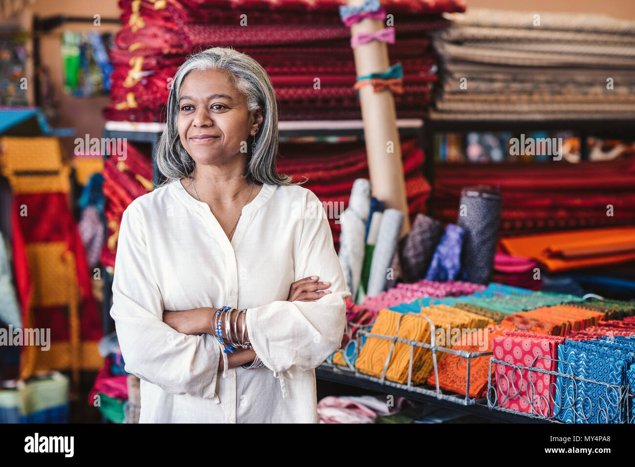 Lächelnd reife Stoff shop besitzer stehen mit ihren Armen durch Regale voll von bunten Textilien und coths gekreuzt umgeben Stockfoto