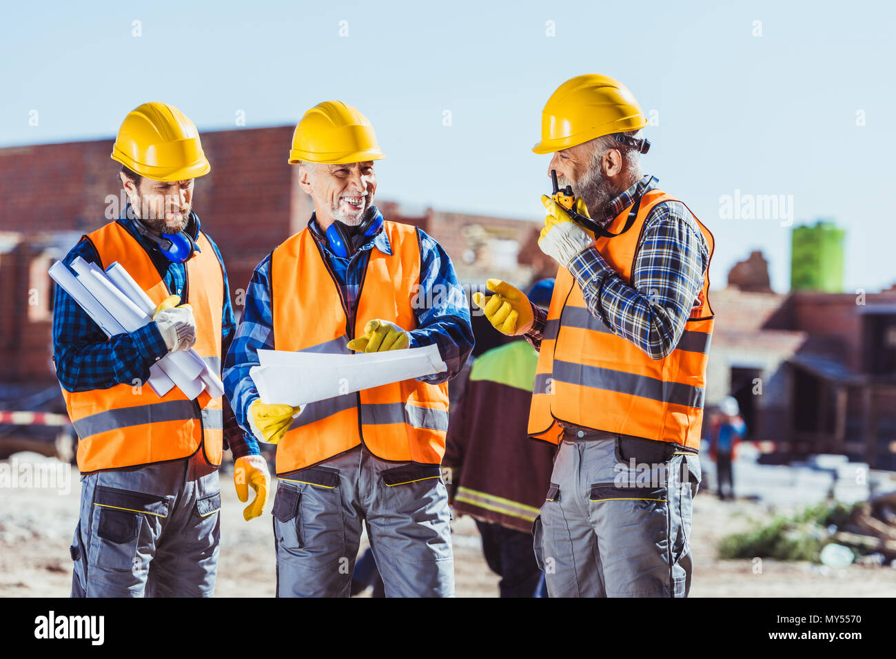 Drei Arbeiter in hardhats Prüfung Baupläne und Sprechen auf tragbaren Radio auf der Baustelle Stockfoto