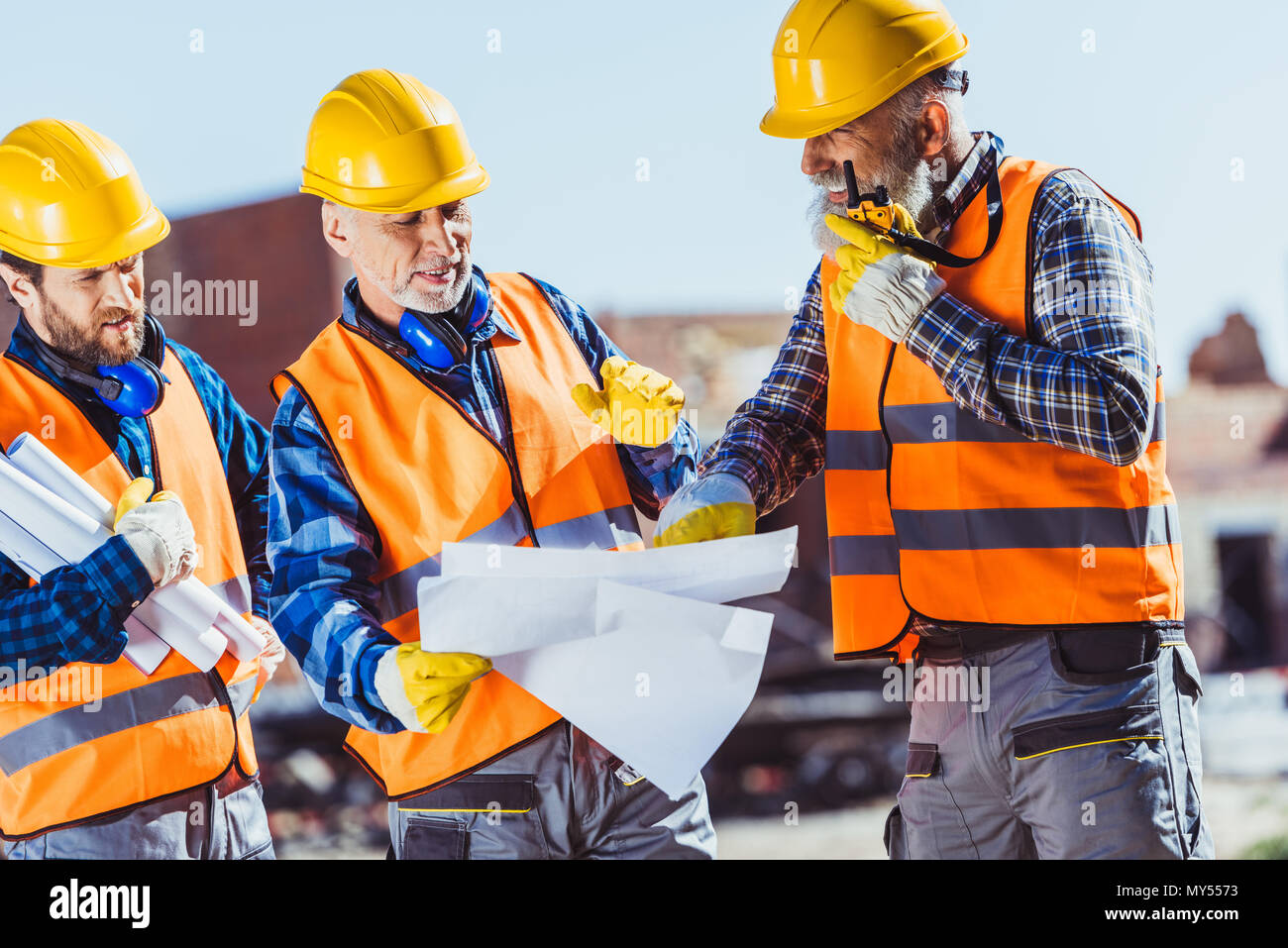 Drei Arbeiter in Uniform Prüfung Baupläne und Sprechen auf tragbaren Radio auf der Baustelle Stockfoto